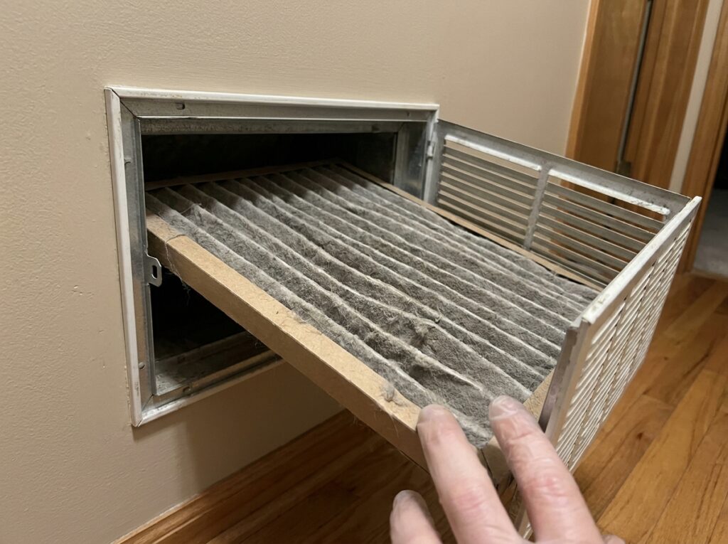 A hand holds open a hinged wall vent grille, revealing a pleated air filter in the opening that is thickly coated with gray dust and lint.